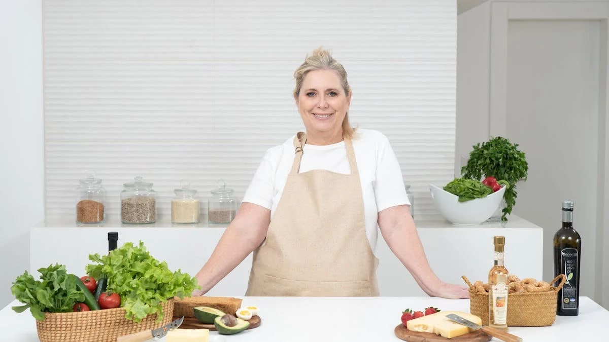 A mature woman in an apron stands at a kitchen counter with fresh produce and cheese, ready for cooking.